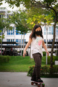 Young girl wearing reusable black face mask skateboarding in the park.