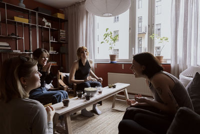 Happy female friends enjoying snacks and drinks while sitting in apartment