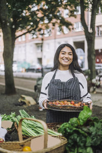 Portrait of young female vendor holding vegetable tray near stall at farmer's market