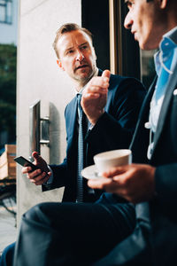 Businessman looking at male colleague talking while holding coffee cup at cafe