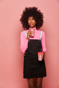 Happy black woman with disposable cup of drink standing against pink background