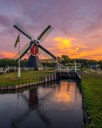 Traditional windmill by river against cloudy sky during sunset