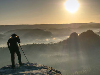 Lady photographer tourist with camera shoots sunrise while standing at tripod on top of mountain.
