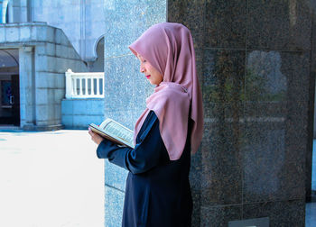 Young woman wearing hijab while reading holy book at mosque