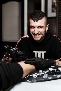 Stylish happy man with piercing looking at camera while using tattoo machine to make tattoo on leg of crop anonymous customer during work in salon