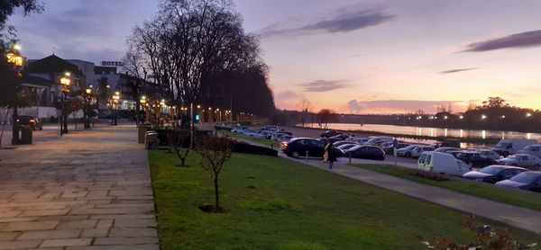 Cars on street by illuminated city against sky at sunset