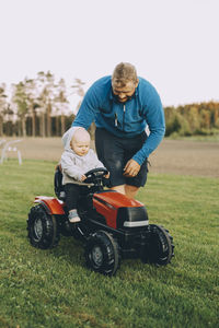 Father giving ride to son sitting in toy tractor at farm