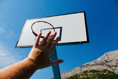 Low angle view of person hand against basketball hoop and blue sky