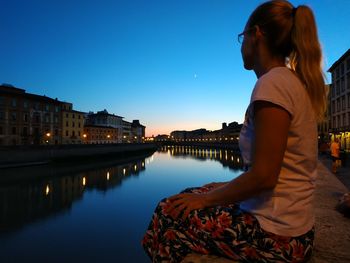 Side view of woman sitting by canal in city against sky