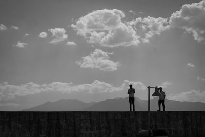 People standing on retaining wall against mountains and sky