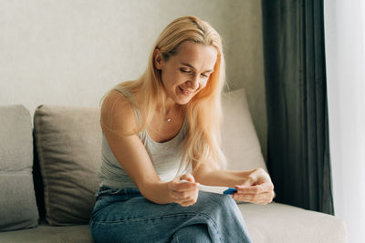 Happy smiling woman holding pregnancy test while sitting on sofa at home.