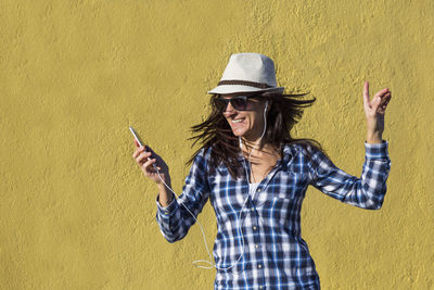 Young woman listening music while standing against yellow wall