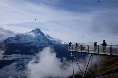 People on footbridge against snow covered mountains and sky