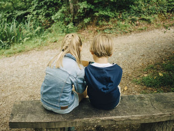 Rear view of siblings sitting on bench by footpath in forest