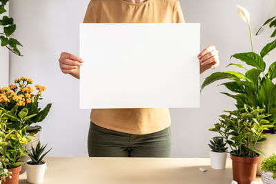 Midsection of woman holding blank billboard standing among house plants in room