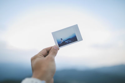 Cropped hand of person holding photograph against sky