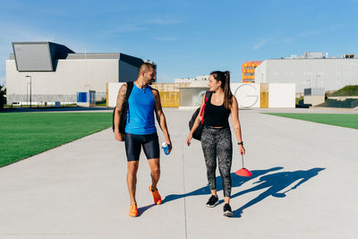 Full body of cheerful athletic man and woman in sportswear with backpacks walking on paved path and talking after fitness workout together in summer day in city