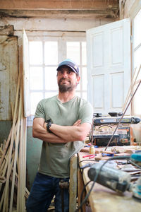 Self assured male woodworker with folded arms and beard looking at camera against instruments in workroom