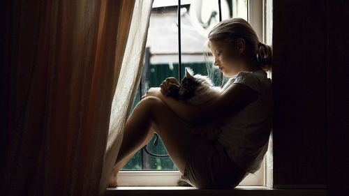 Side view of girl sitting with cat on window sill