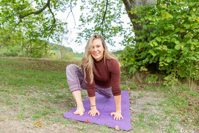 Smiling young woman sitting in a yoga pose in a park on a yoga mat with a tree on a background