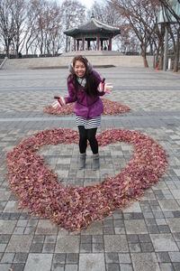 Full length portrait of smiling young woman standing amidst heart shape fallen leaves on footpath