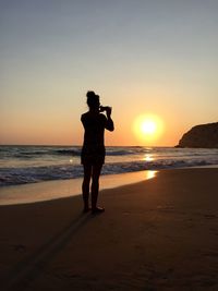 Rear view of silhouette woman photographing while standing at beach against clear sky during sunset