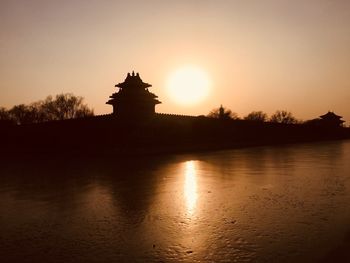 Silhouette building by lake against sky during sunset