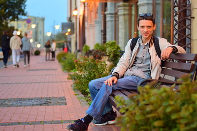 Full length portrait of smiling man sitting on bench in city