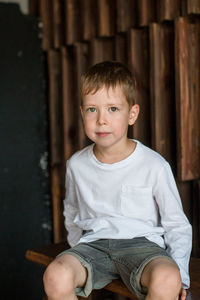 Portrait of a little blond boy in a white t-shirt sits against the background of a wooden wall 