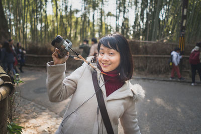 Portrait of smiling young woman holding camera while standing against trees