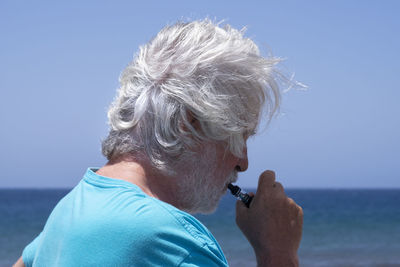 Side view of senior man smoking electronic cigarette against sky
