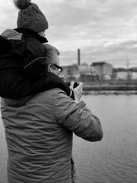 Rear view of father carrying daughter while standing by river against cloudy sky