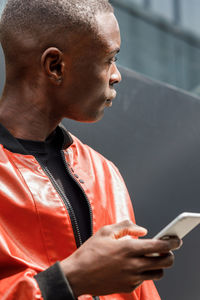 Side view of concentrated young african american guy in trendy red leather jacket messaging on mobile phone while leaning on wall on city street