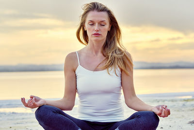 Woman meditating while sitting by lake against sky during sunset