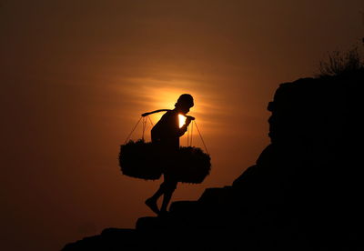 Silhouette man with baskets carrying on shoulders against sky during sunset