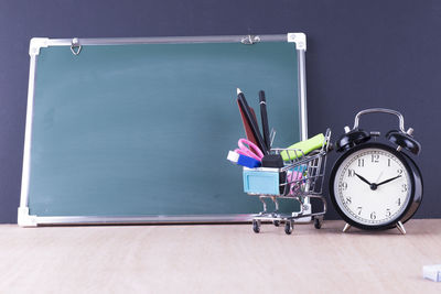 Shopping cart with school supplies by blackboard and alarm clock against wall