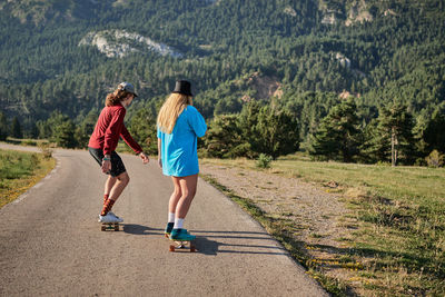 Back view of couple of hipsters in fancy wear riding skateboards together along asphalt road in mountainous area in summer