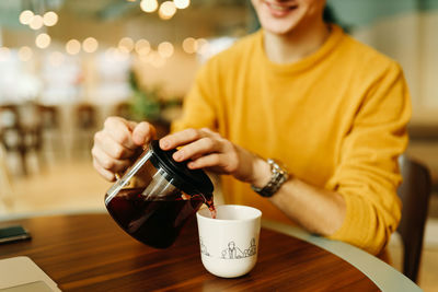 A smiling man in a bright yellow sweater pours tea coffee into a mug from a teapot sitting in a cafe