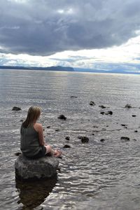 Rear view of woman sitting on rock at shore against cloudy sky