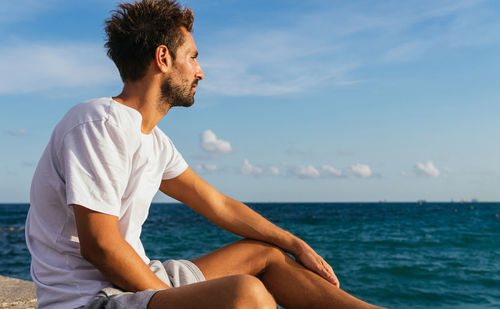 Hispanic male athlete in activewear sitting on embankment against cloudy blue sky and admiring sea during break in fitness training on summer day
