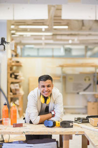 Portrait of smiling teenage trainee leaning on workbench at workshop