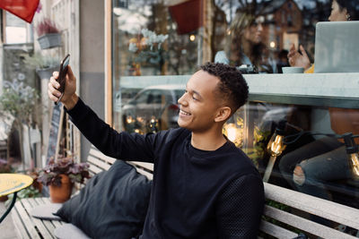Smiling young man sitting on bench taking selfie against cafe window