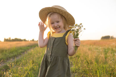 A little blonde girl in a straw hat walks in a field with a bouquet of daisies. 