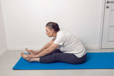 Elderly woman does yoga on a blue rug in a white room at home