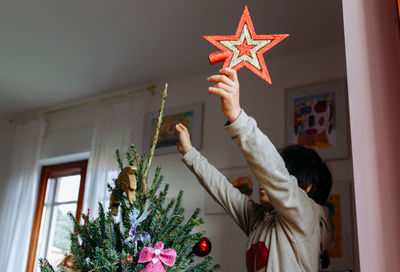 Low angle view of child putting red star on top of christmas tree at home