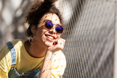 Modern cheerful millennial hispanic female with curly hair wearing yellow sweatshirt with denim overalls and trendy sunglasses and earrings sitting leaning on hand near mesh fence in sunlight