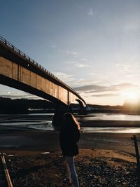 Rear view of woman standing on shore by bridge against sky