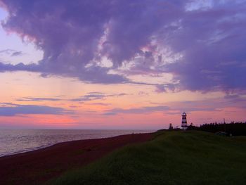 Lighthouse on hill by sea against cloudy sky during sunset