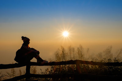 Silhouette woman sitting on fence against bright sun during sunrise