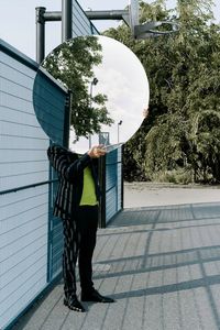 Man holding mirror while standing on footpath against trees